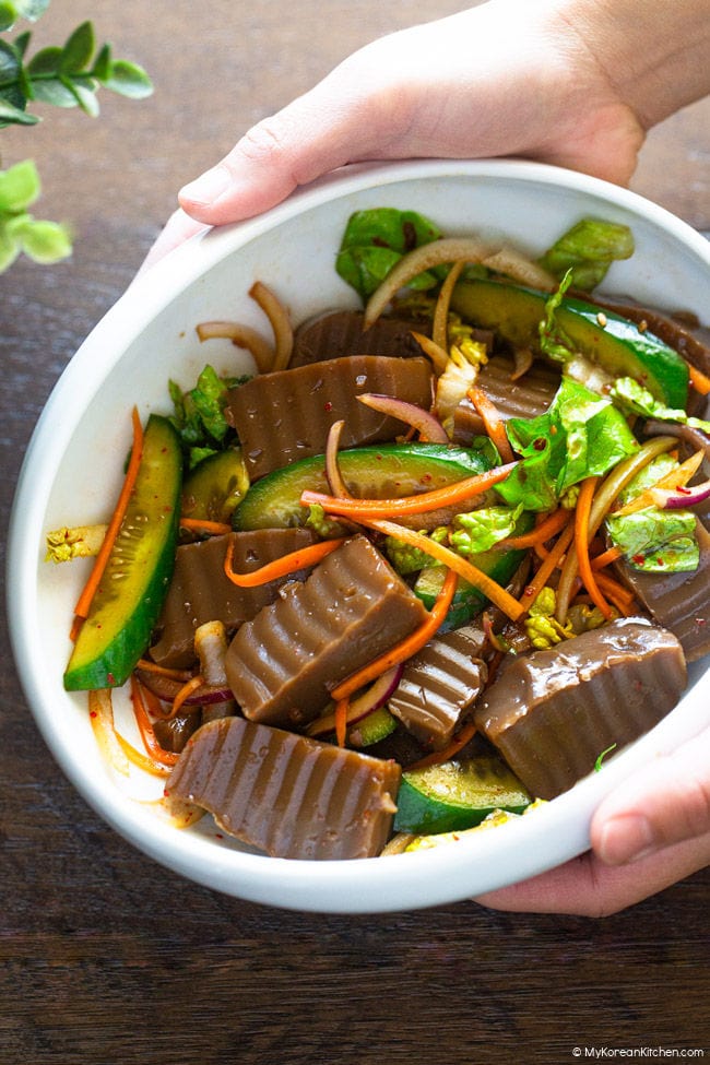 Hands holding a bowl of seasoned acorn jelly salad, placing it on a wooden table.