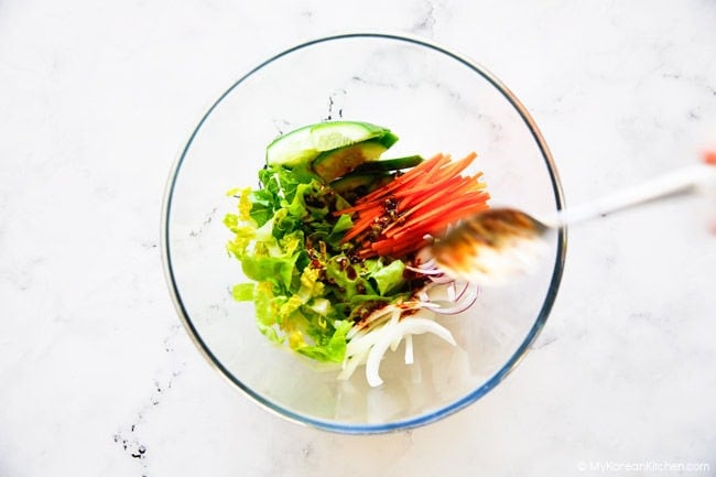 Adding salad sauce over fresh vegetables in a clear mixing bowl.