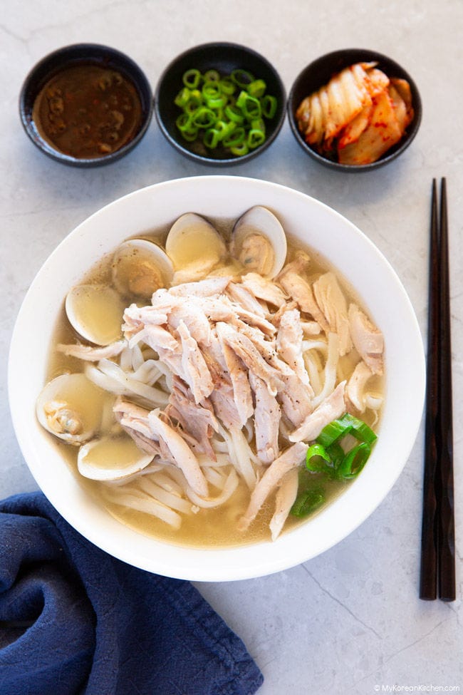 Dak kalguksu is nicely presented in a white bowl, accompanied by chopped sauce, green onions, and kimchi.