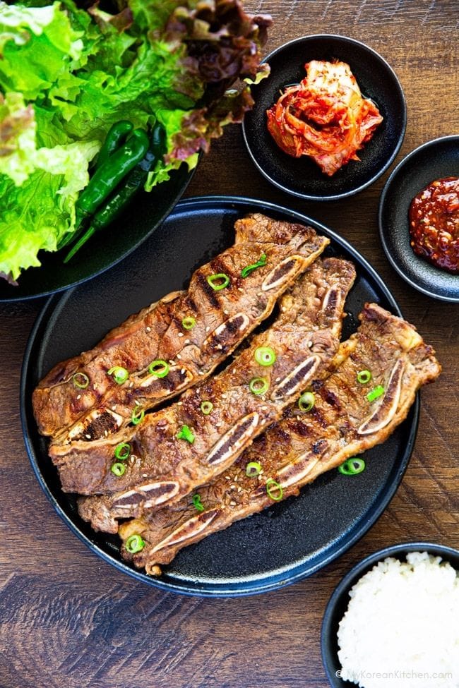 Grilled LA galbi on a black plate set on a wooden board, served with lettuce, kimchi, ssamjang, and a bowl of white rice