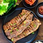 Grilled LA galbi on a black plate set on a wooden board, served with lettuce, kimchi, ssamjang, and a bowl of white rice