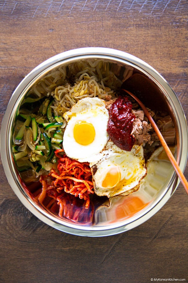 bibimbap ingredients in a large stainless steel bowl, including a sunny side egg, zucchini, spicy radish salad, bean sprout salad, and gochujang.
