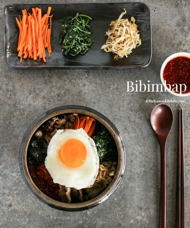 Bibimbap topped with an egg. Served with chopsticks and a spoon-- Korean rice bowl recipe