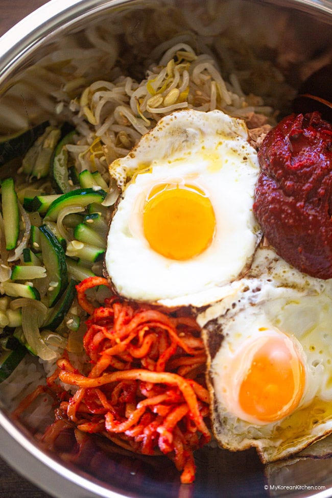 Close-up photo of bibimbap ingredients in a large stainless steel bowl, including two sunny-side eggs, zucchini, spicy radish salad, bean sprout salad, and gochujang