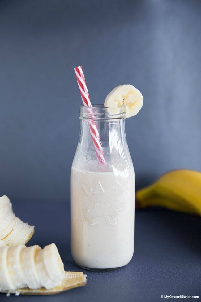 A jar of banana milk with a straw. Sliced banana sitting next to the Korean drink. 