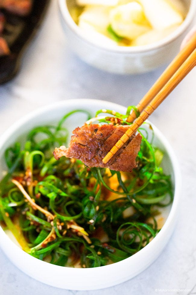 Holding a cooked Korean BBQ pork with a green onion salad with a pair of chopsticks.