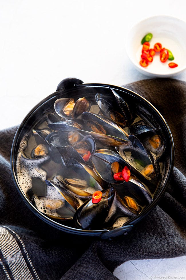 Boiled mussel soup in a black pot. A small bowl of sliced chilies is on the top right corner.
