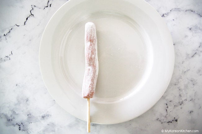 A sausage on a stick is covered with flour and placed on a white plate.