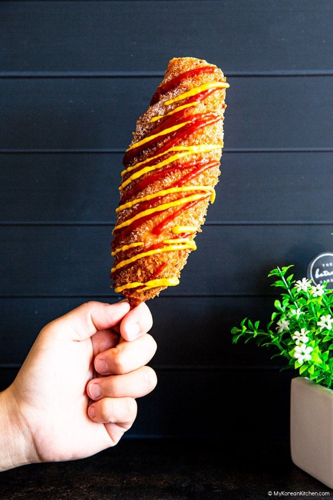 Holding a Korean corn dog in one's hand in front of a black wooden board background.