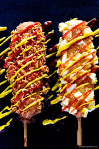 Two Korean corn dogs lay next to each other on a black background board, with a splash of mustard and ketchup sauce over them.