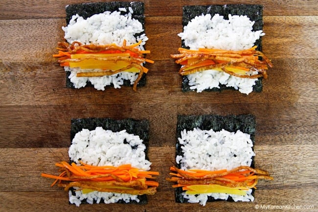 Four mini kimbap are arranged on a wooden board ready to be rolled up.