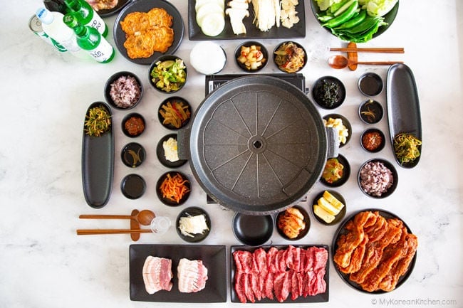 A horizontal photo of a Korean BBQ table set up with various side dishes and meat.