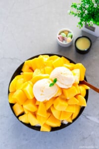 A black bowl is filled with a pile of cut mangoes, topped with two scoops of ice cream.