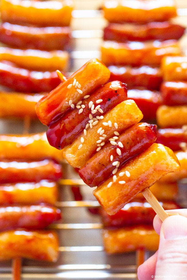Holding one Sotteok skewer in one hand, with more Sotteok skewers visible on a plate in the background.