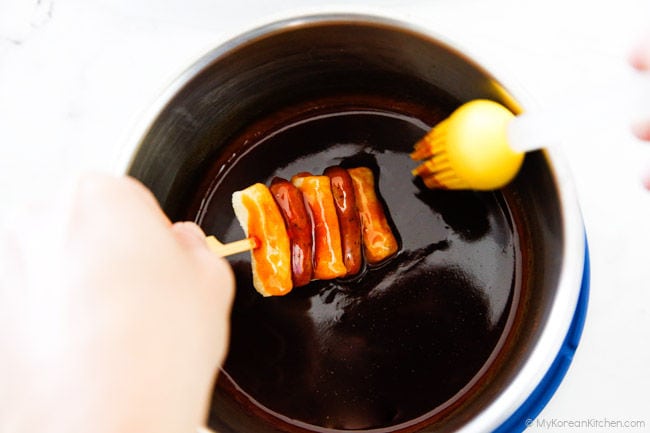 Holding the sotteok skewers in one hand, then basting them with sotteok sauce using a yellow silicone brush.