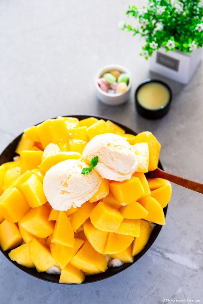 A black bowl filled with a pile of sliced mangoes, topped with two scoops of ice cream, is positioned towards the left side of the photo.