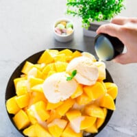 Glazing sweetened condensed milk over a pile of sliced mangoes in a black bowl