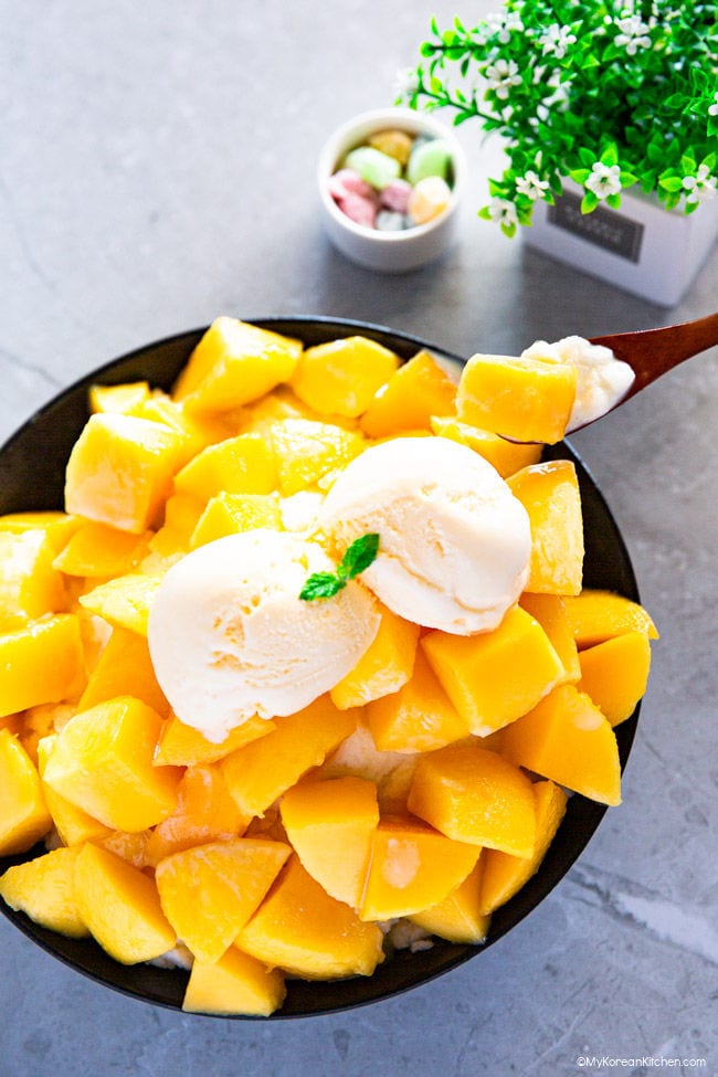 A black bowl is filled with a pile of cut mangoes, topped with two scoops of ice cream. A brown wooden spoon is used to scoop up some of the mangoes and shaved ice.