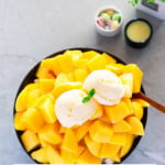 A black bowl is filled with a pile of cut mangoes, topped with two scoops of ice cream, with a text overlay saying “Mango Bingsu”.