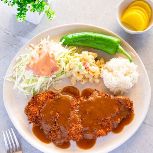 A plate filled with donkatsu covered in sauce, shredded cabbage topped with a ketchup-mayo blend, a side of green chili, macaroni salad, and a scoop of white rice.