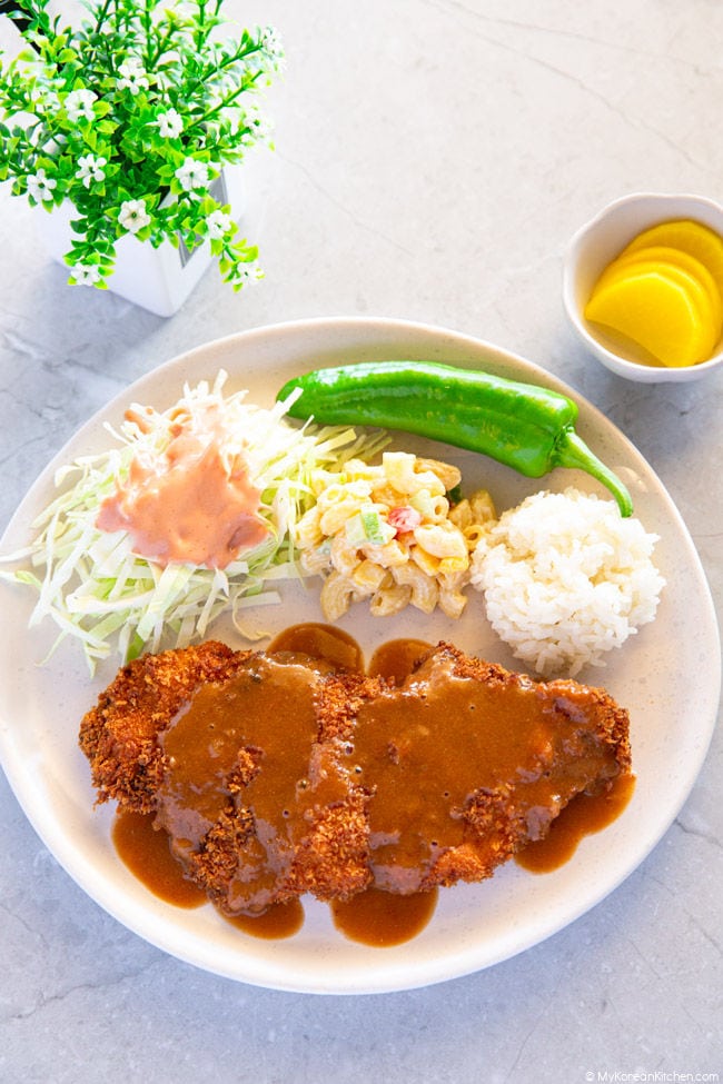 A plate filled with donkatsu covered in sauce, shredded cabbage topped with a ketchup-mayo blend, a side of green chili, macaroni salad, and a scoop of white rice.