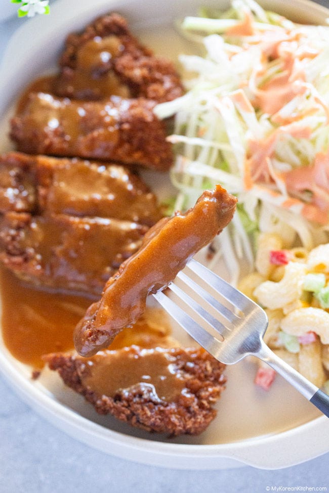 Close-up photo of a slice of donkatsu on a fork, coated with sauce, over a plate.