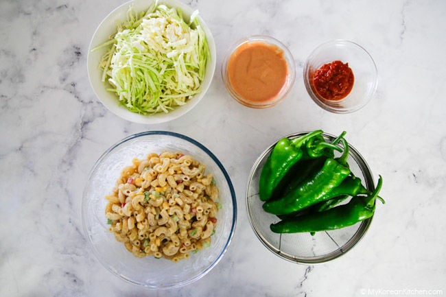 Shredded cabbage, ketchup-mayo dressing, ssamjang, a large bowl of macaroni salad, and green chilies on a white marble backdrop.