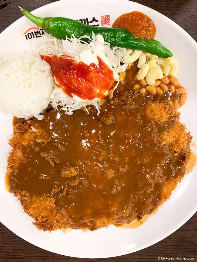 Jumbo sized donkatsu served on a white plate, accompanied by rice, shredded cabbage, a green chili, macaroni salad, and baked beans.