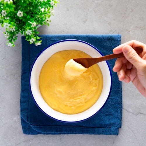 A hand holding a wooden spoon, scooping vanilla custard from a serving bowl.