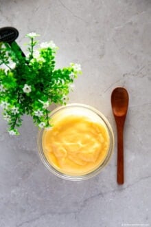 Custard in a small glass container, with a small wooden spoon on the side. In the background, there are small green plants.