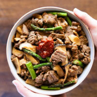 Holding a bowl of cooked Korean BBQ pork with vegetables in two hands.