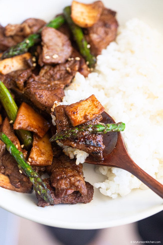 A wooden spoon filled with rice, BBQ meat, mushrooms, and asparagus, resting on a bed of white rice.