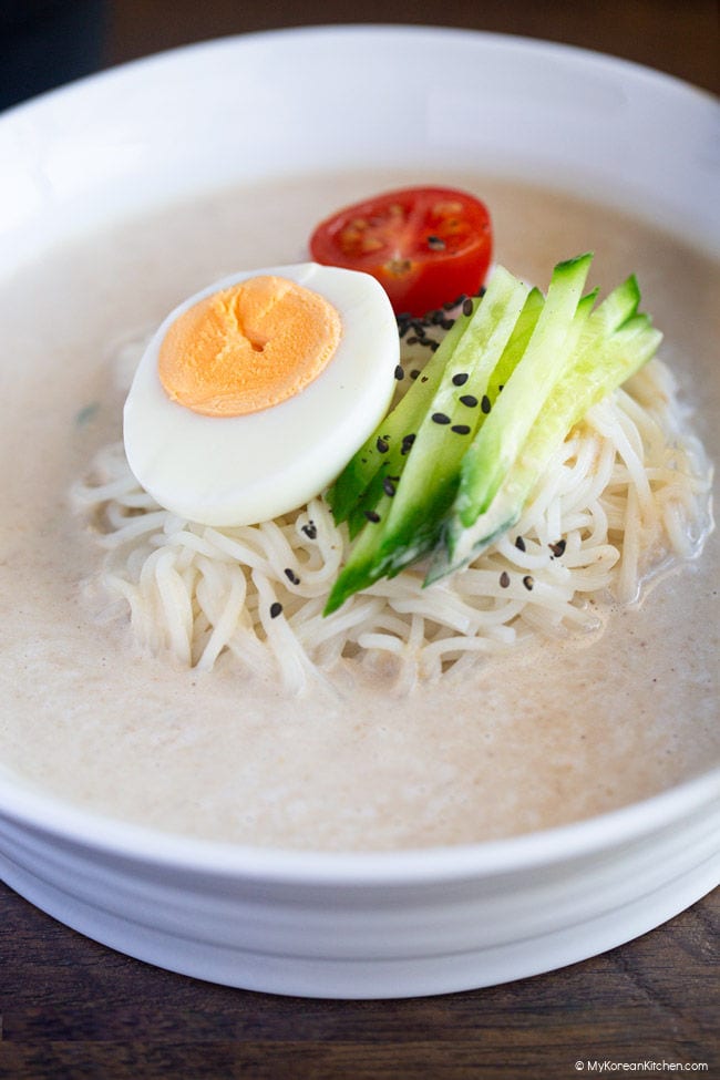 Close-up photo of noodles in a white bowl, topped with halved cherry tomatoes, a halved boiled egg, and julienned cucumbers.