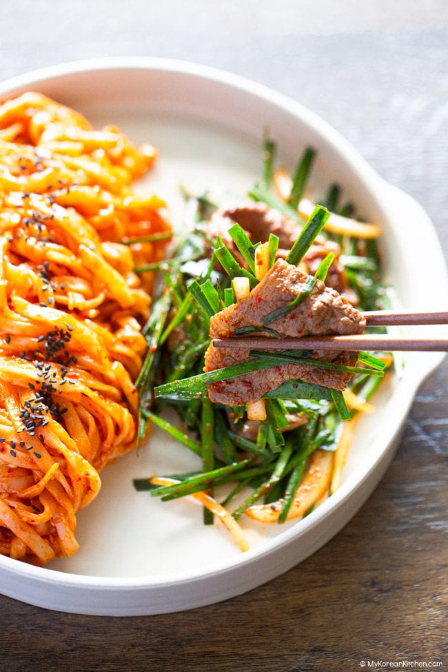 Chopsticks holding brisket salad, with spicy knife-cut noodles on a plate beside it.