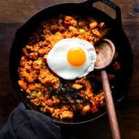 Radish kimchi and spam fried rice served in a cast iron skillet with a sunny-side-up egg on top and a wooden ladle on the side.