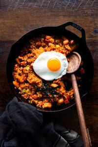 Radish kimchi and spam fried rice served in a cast iron skillet with a sunny-side-up egg on top and a wooden ladle on the side.