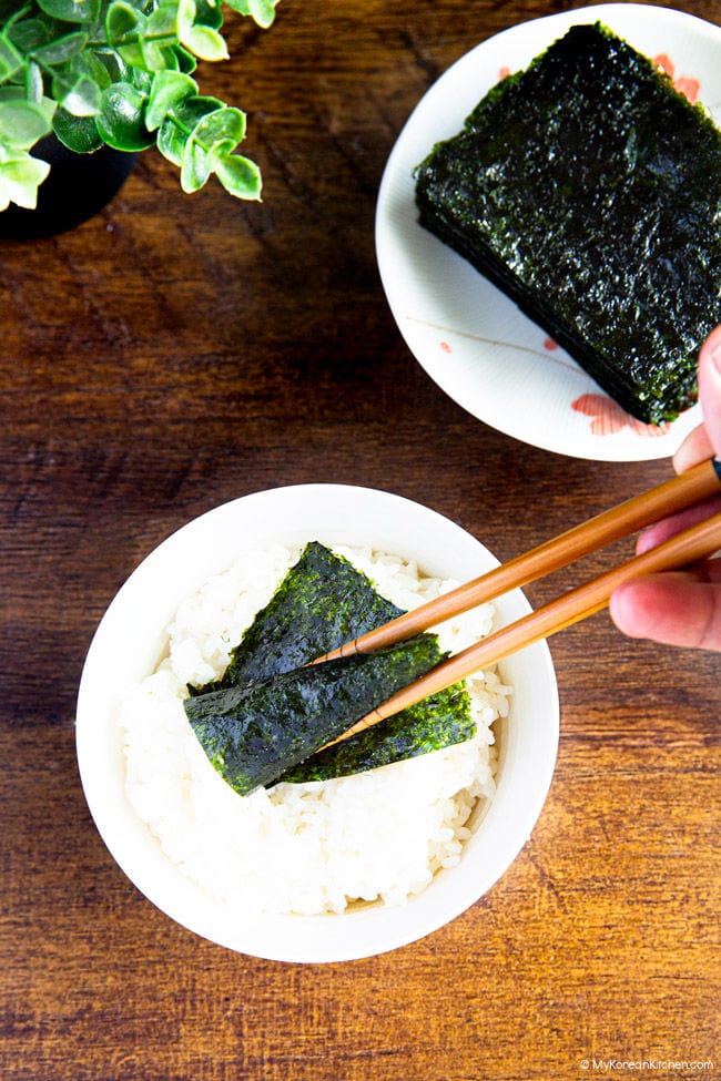 Wrapping cooked rice with roasted seaweed using wooden chopsticks over a bowl of rice.