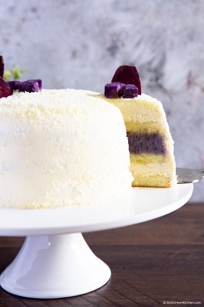 Front shot of a Korean sweet potato cake on a white cake stand with a slice being taken out.