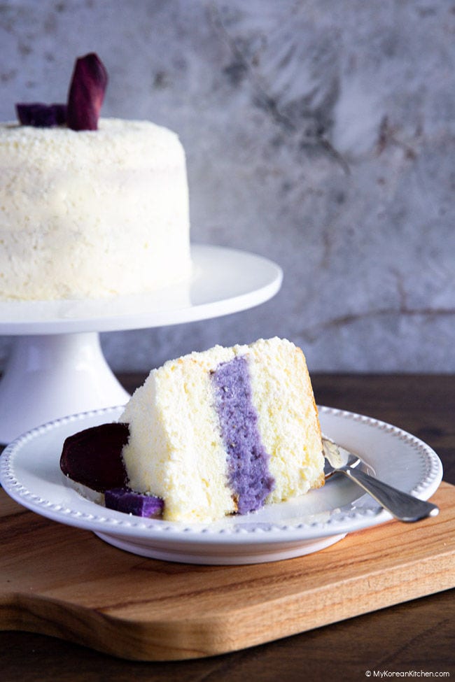 A slice of Korean sweet potato cake on a white serving plate, with the remaining cake on a cake stand in the background.