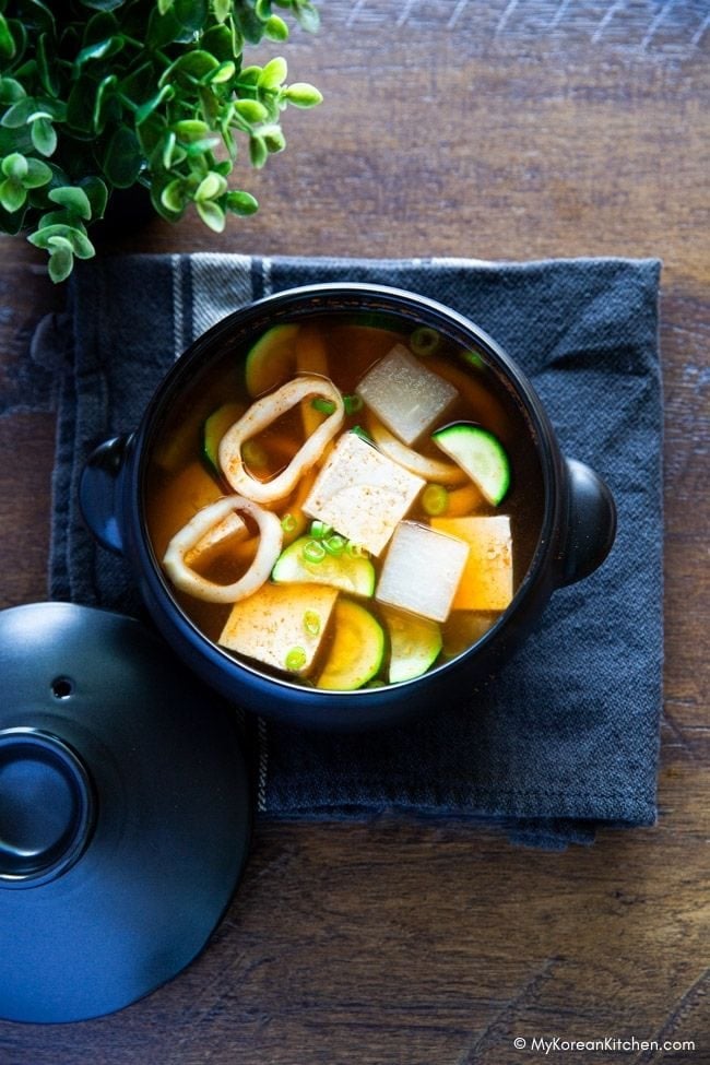 A black pot filled with Korean squid soup containing tofu, vegetables, and squid rings, placed on a wooden bench.
