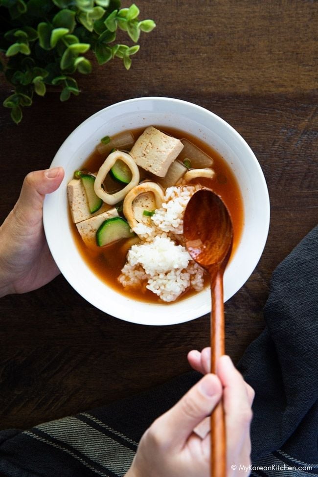A person mixing rice into a bowl of squid soup with a wooden spoon.