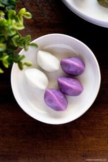 Purple and white songpyeon served on a small white plate on a wooden board