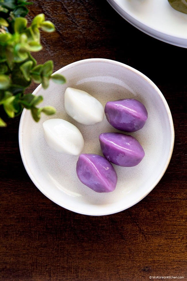 Purple and white songpyeon served on a small white plate on a wooden board