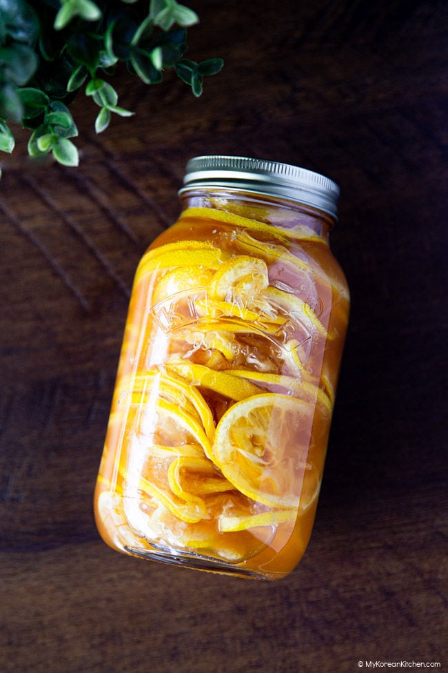 Angled view of a glass jar filled with Korean lemon ginger syrup, showing vibrant lemon slices in golden honey syrup on a rustic wooden background.