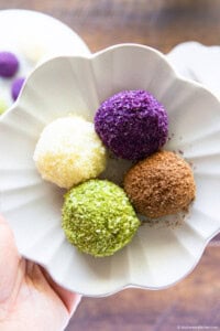 Hand holding a small flower-shaped plate with four colorful sweet rice balls.