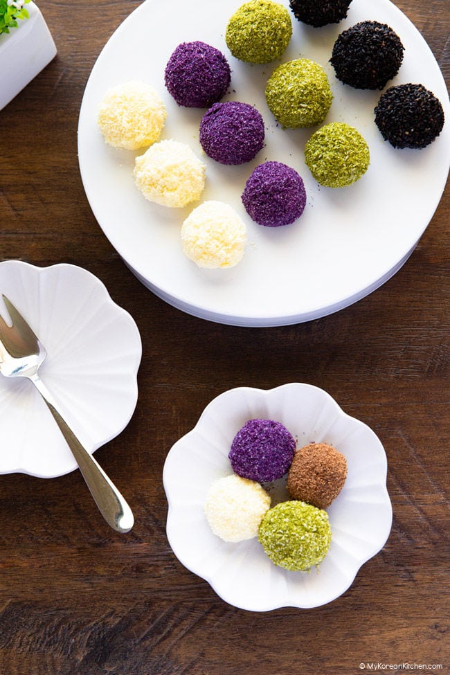 Assorted gyeongdan coated in various toppings on a white platform plate, with four more rice balls on a small flower-shaped dish.