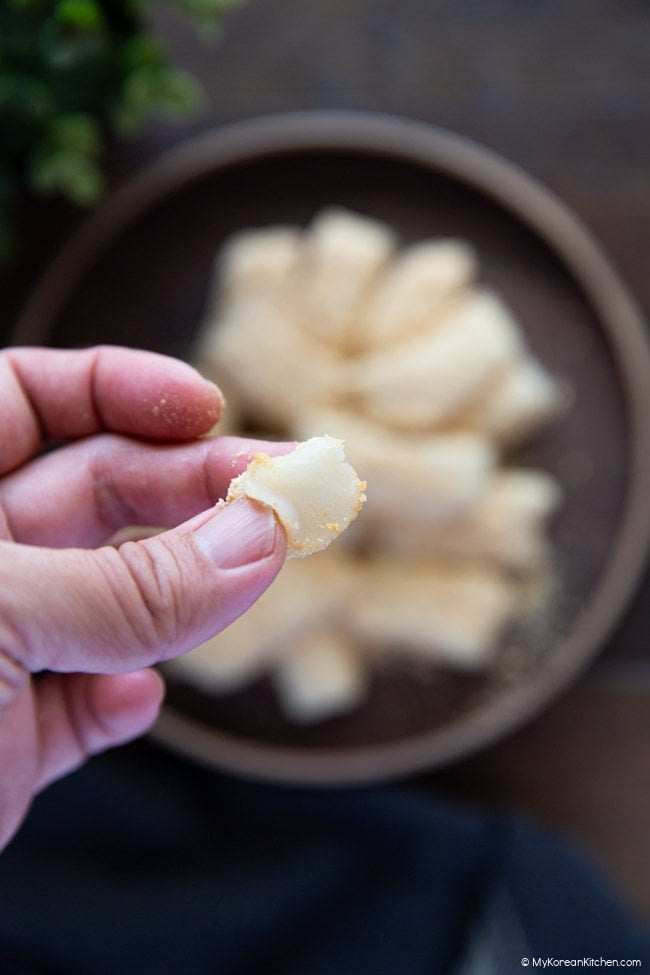 Half-eaten injeolmi Korean rice cake held between fingers with more injeolmi on a wooden plate in the background