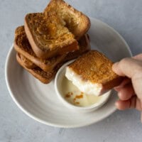 golden butter tteok stacked in the background, with a hand holding a broken piece dipped in sweetened condensed milk