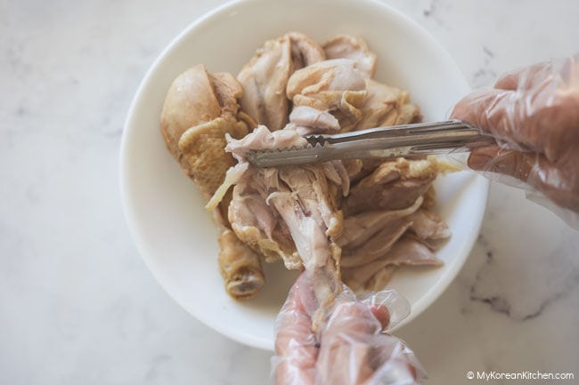 Cooked chicken meat being separated with tongs and a gloved hand