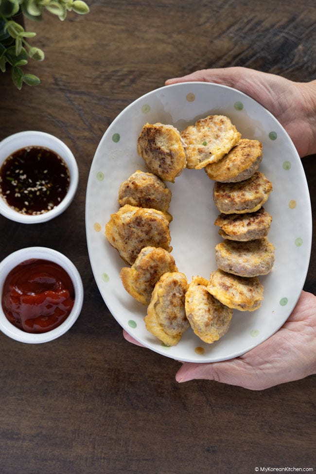 A plate of Korean meat patties held in two hands, with two dipping sauces on the side.